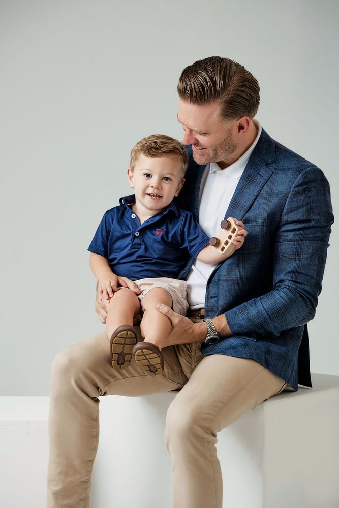Father in a blue plaid blazer with his smiling toddler son on his lap holding a wooden toy car, studio portrait
