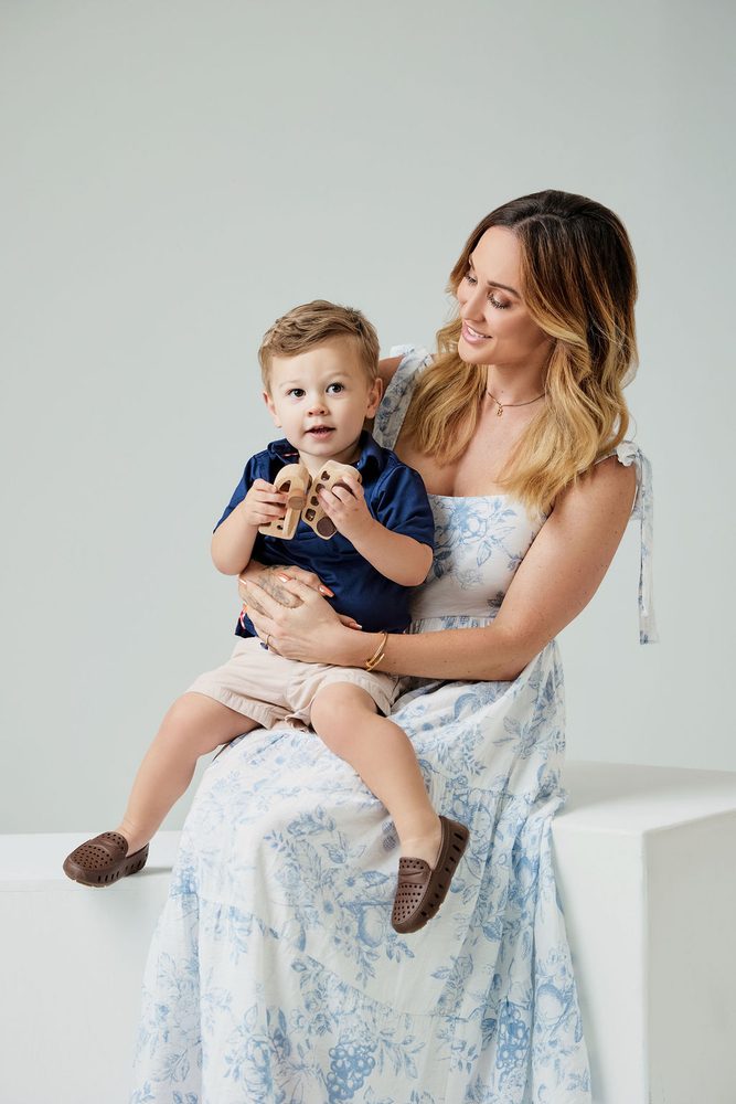 Mother in a blue floral sundress with her toddler son on her lap holding wooden toys, seated on a white block