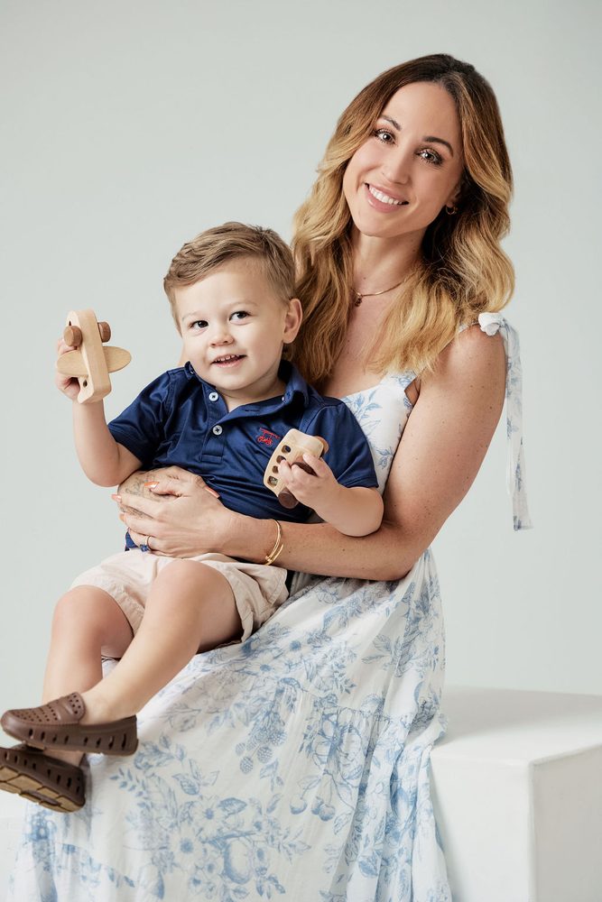 Smiling mother on a white studio block holding her toddler, who shows off a wooden toy airplane
