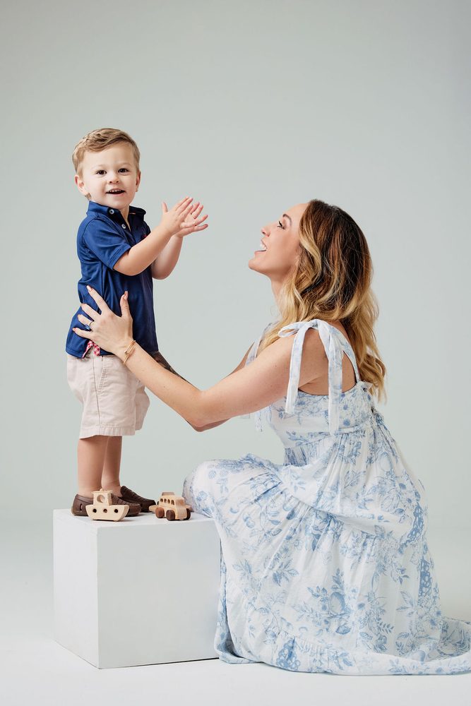 Toddler standing on a white block clapping as his kneeling mother in a blue floral dress steadies him, toys at his feet