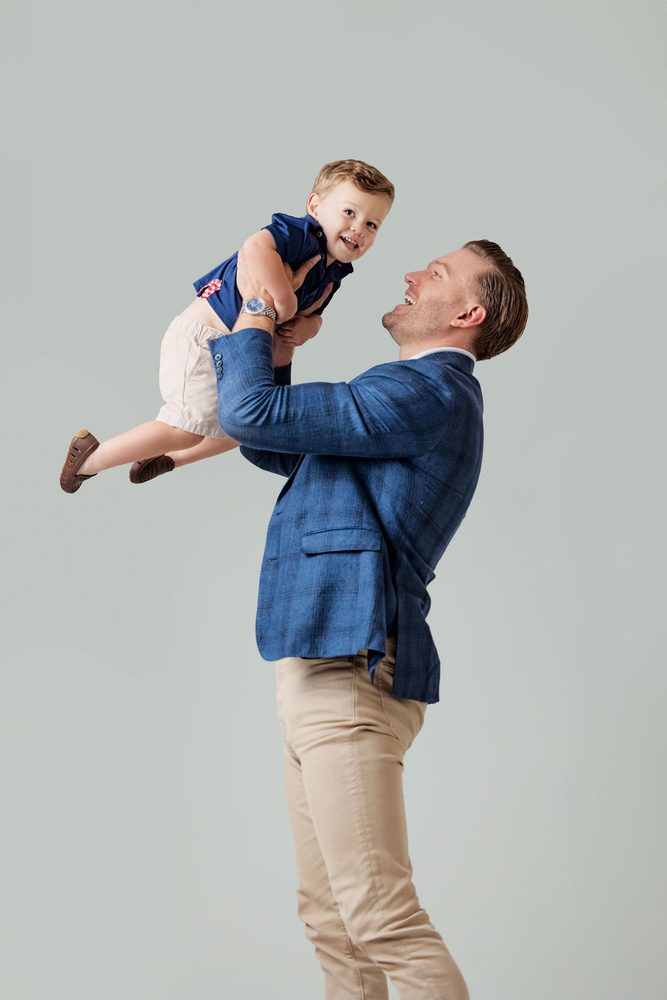 Father in a blue blazer lifting his laughing toddler son high in the air against a light grey studio backdrop