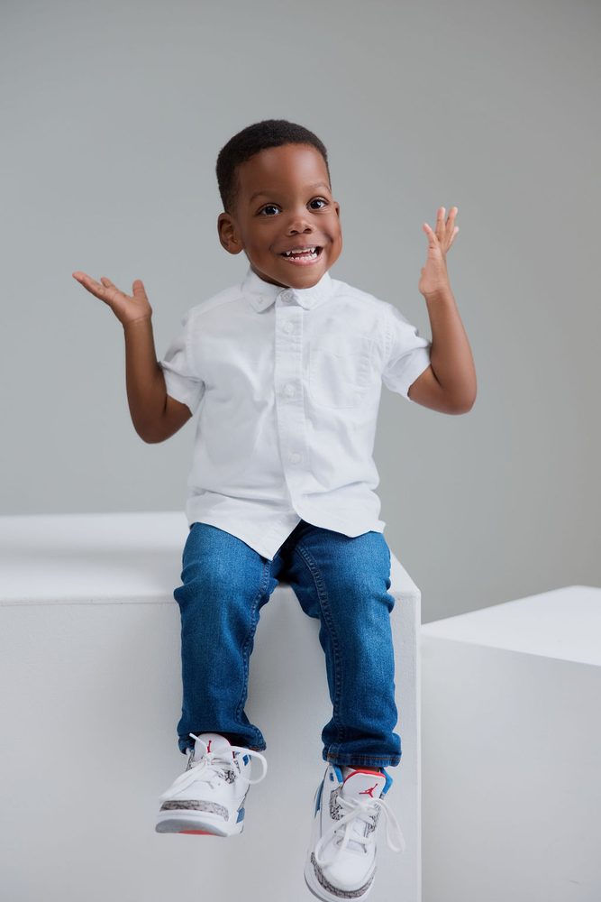 Excited young boy in a white shirt and jeans sitting on a white studio block with both hands raised