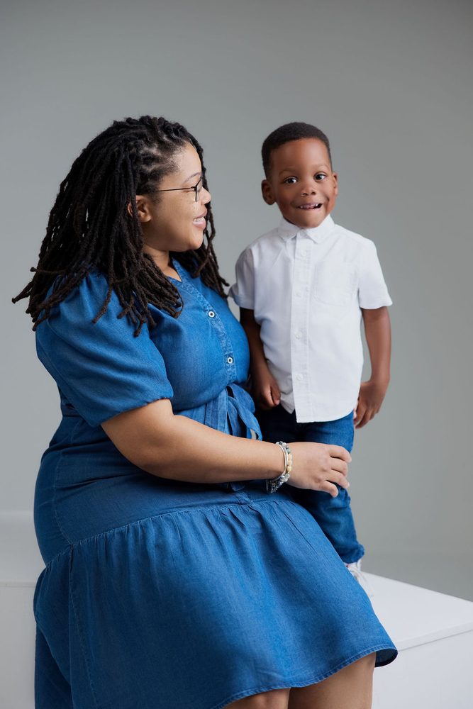 Mother in a denim dress and glasses smiling at her young son in a white shirt standing beside her in the studio