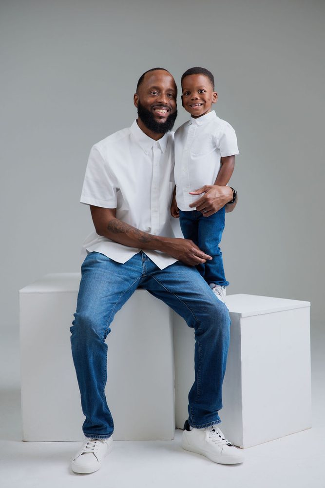 Smiling father in a white shirt and jeans seated on studio blocks with his young son standing beside him