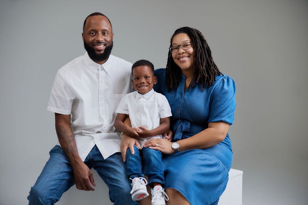 Family of three in white and denim seated together on a studio block, young son on his parents' laps
