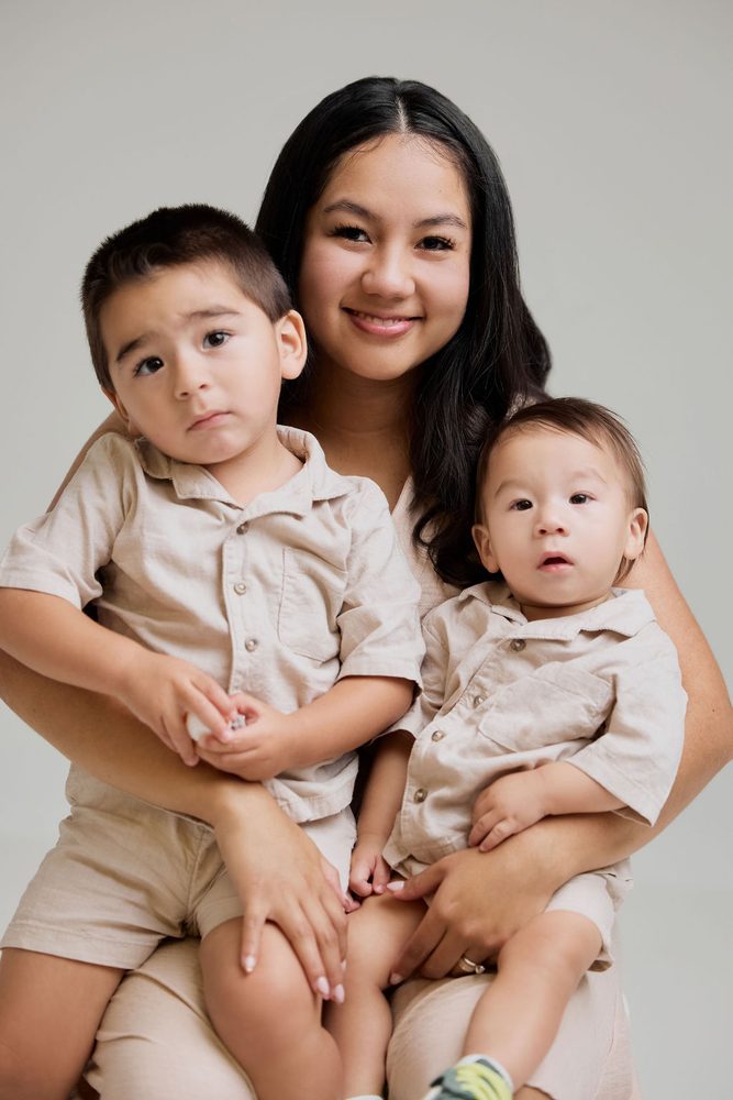 Mother holding her two young sons in matching beige linen outfits, close-up studio portrait on grey