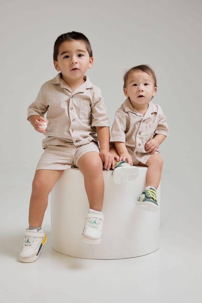 Two young brothers in matching beige linen shirts and shorts sitting on a white round pedestal