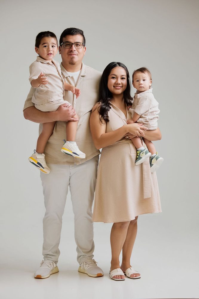 Full-length portrait of parents in neutral tones each holding a young son, light grey studio backdrop