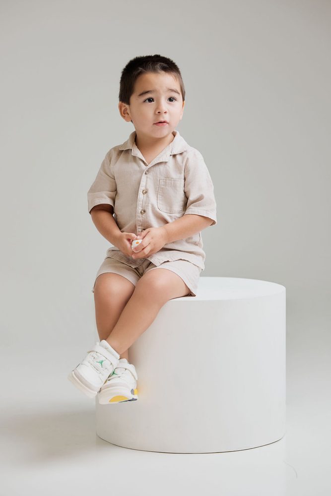 Toddler boy in a beige linen outfit and white sneakers sitting alone on a white round pedestal