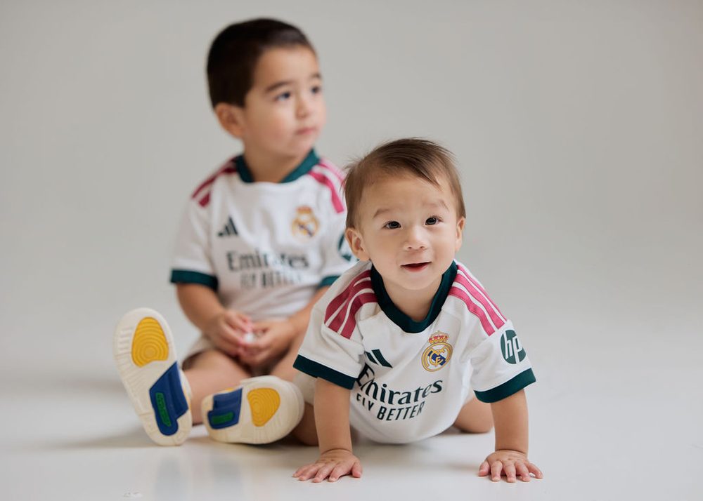 Two young brothers in matching white soccer jerseys, the baby crawling toward the camera on a white studio floor