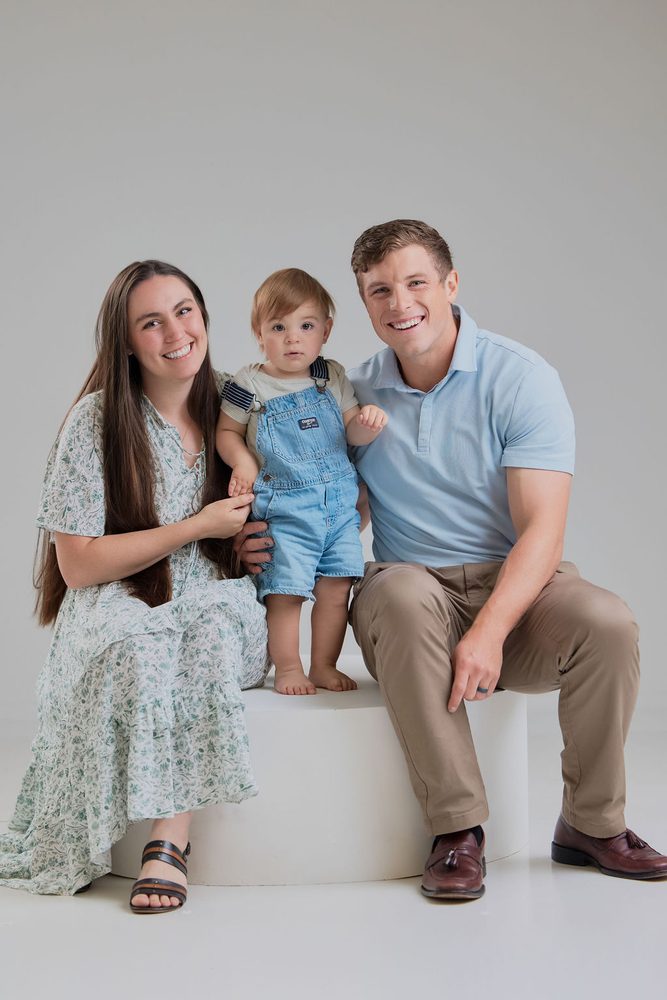 Smiling parents seated on a white pedestal with their baby boy in denim overalls standing between them