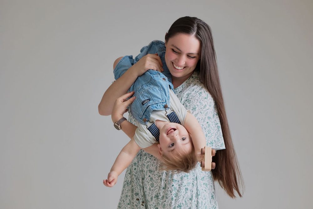 Laughing mother in a green floral dress swinging her baby upside down in denim overalls, grey backdrop