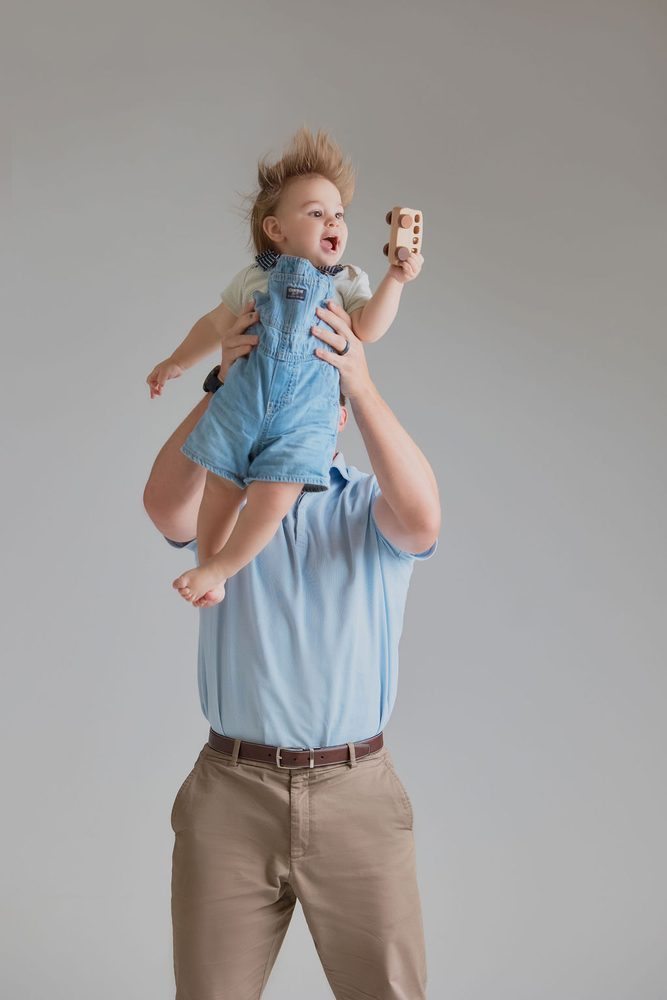 Father in a light blue polo lifting his baby in denim overalls overhead, hair flying, wooden toy in hand