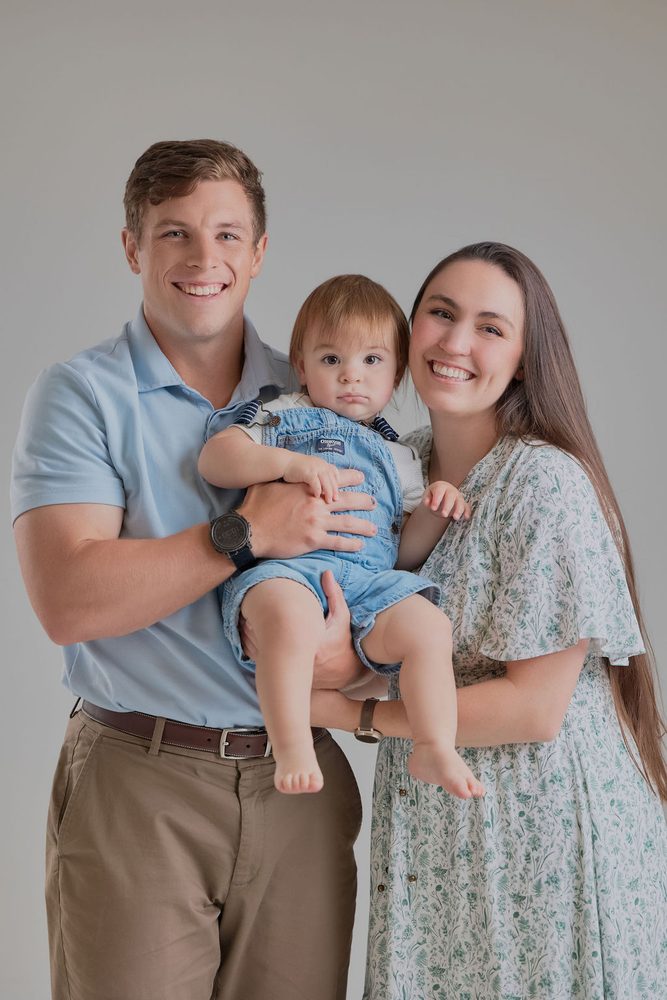 Parents standing close and holding their baby son in denim overalls between them, soft grey studio