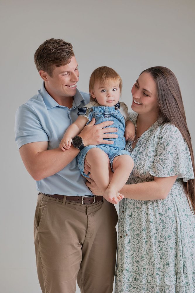 Parents smiling at each other while holding their baby in denim overalls, father in blue polo, mother in florals