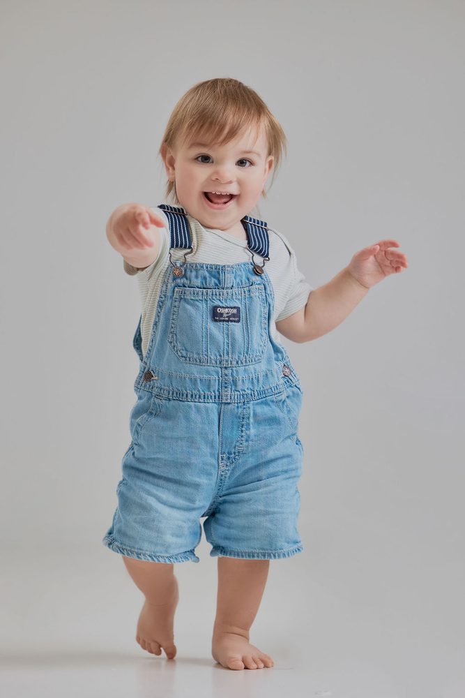 Barefoot baby boy in denim overalls taking happy steps toward the camera, arm outstretched, grey studio