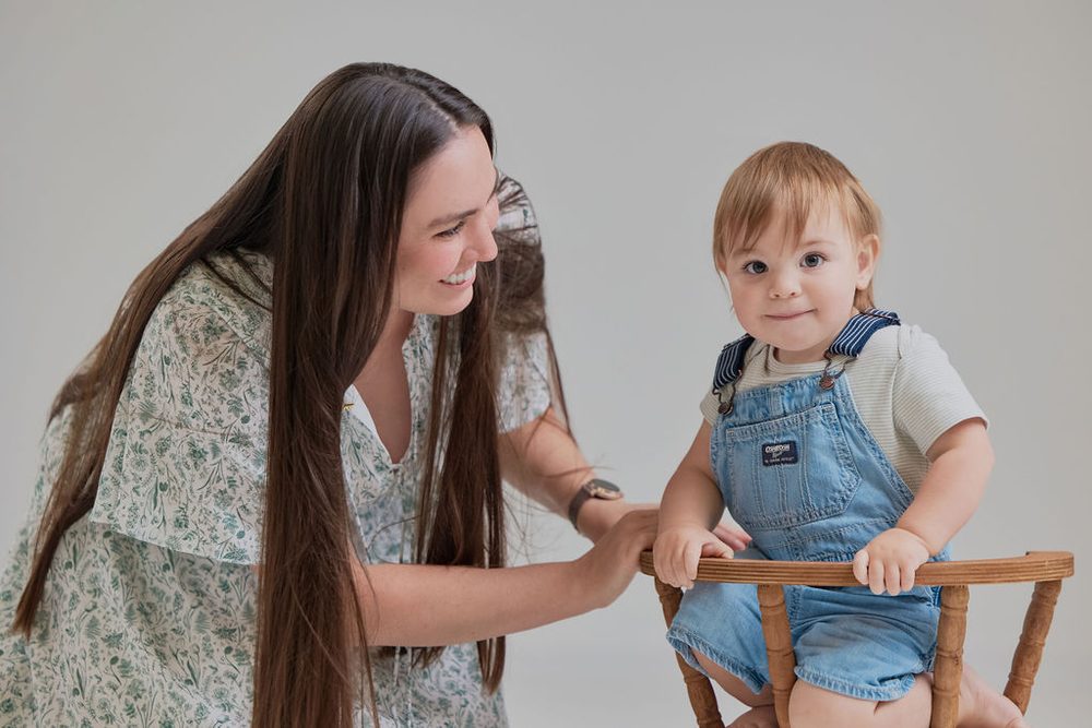 Smiling mother leaning toward her baby son as he stands holding the rail of a small wooden chair