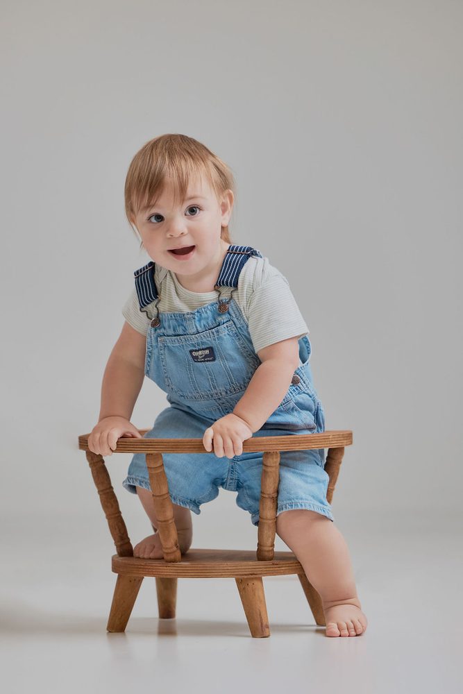 Baby in denim overalls standing and gripping a small wooden stool, looking at the camera on a grey backdrop