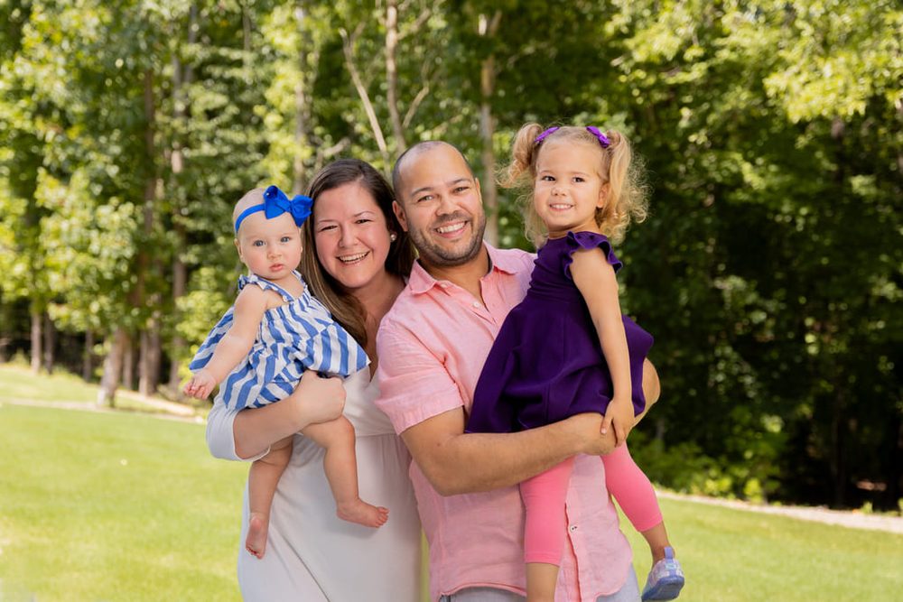 Family of four outdoors on a sunny lawn, parents each holding a young daughter, green trees behind