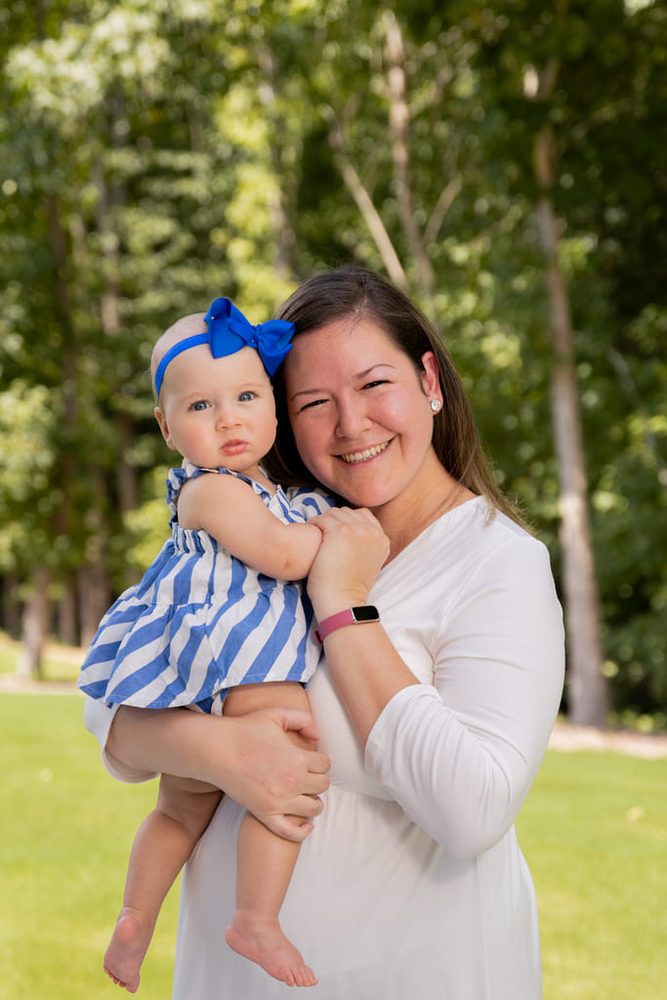Smiling mother in white holding her baby girl in a blue striped dress and big blue bow, outdoors by trees