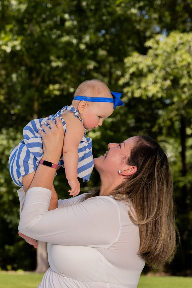 Mother lifting her baby girl in a blue striped dress overhead, smiling up at her against green summer trees