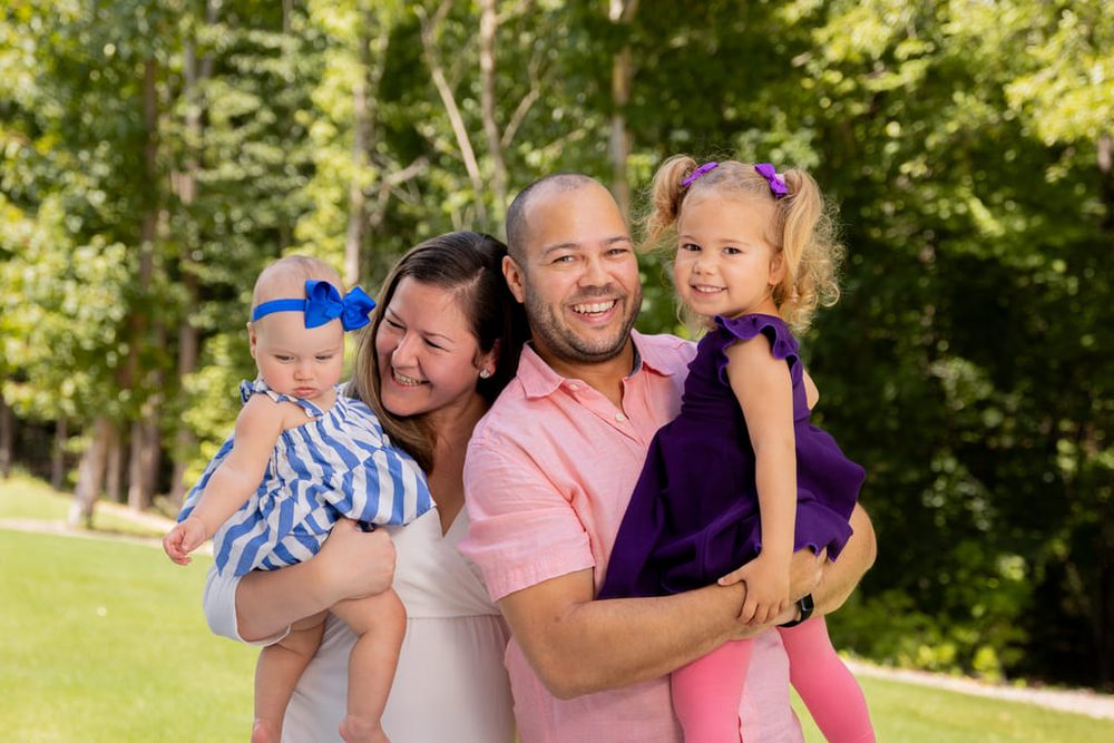 Laughing family of four on a sunny summer lawn, daughters in blue stripes and purple held by their parents