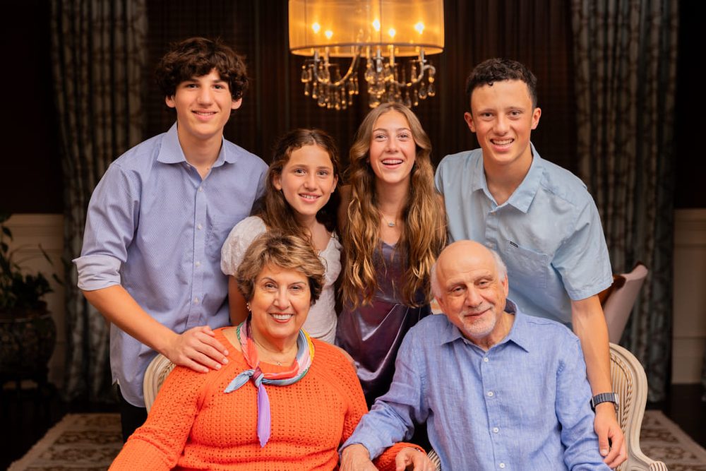 Grandparents seated in front of four smiling grandchildren under a chandelier in an elegant living room