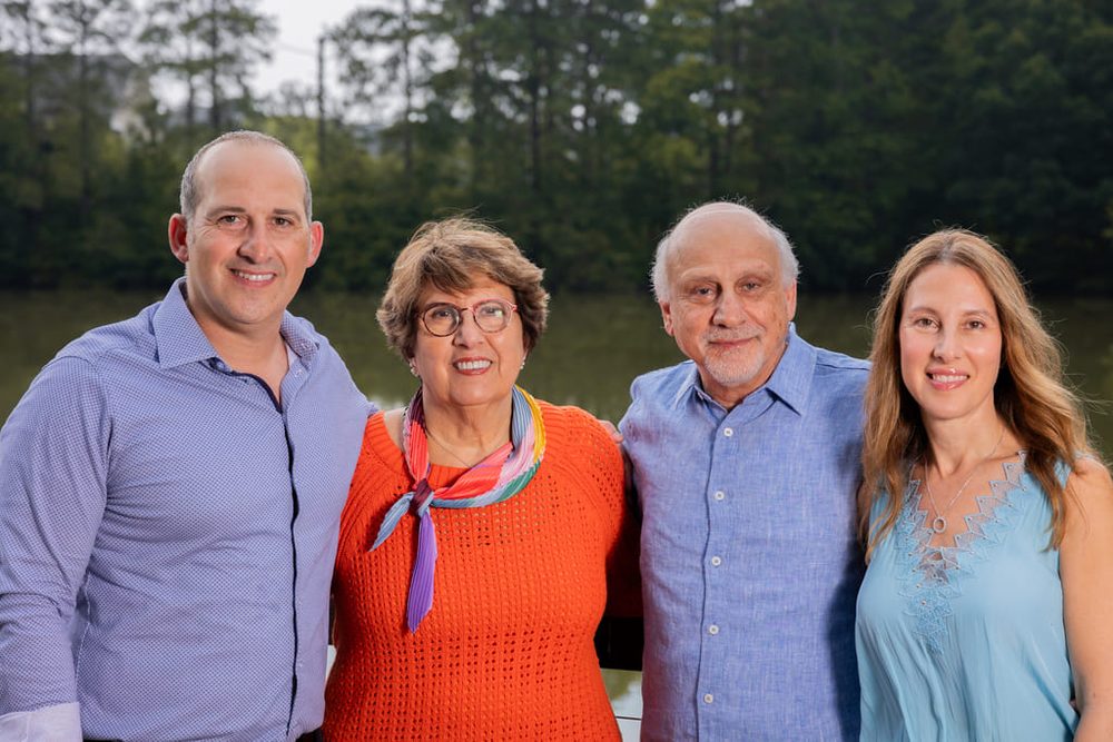 Grandparents standing with two adult children by a lake, smiling, with a wooded shoreline behind