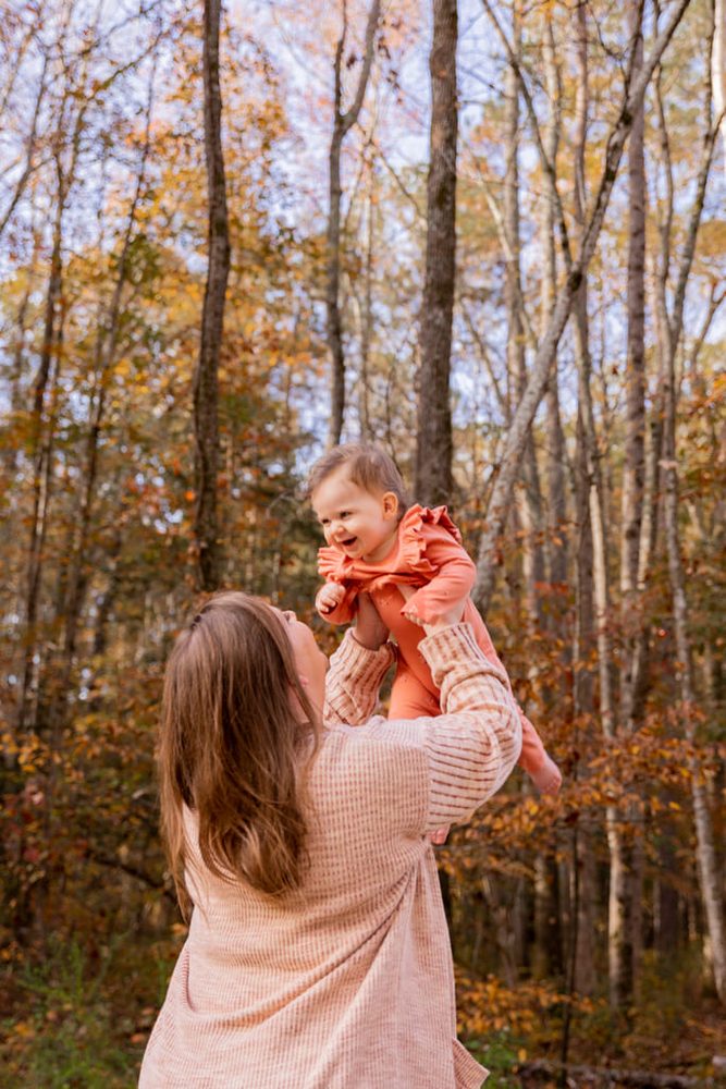 Mother lifting her laughing baby girl in a coral ruffled outfit among autumn trees in the woods