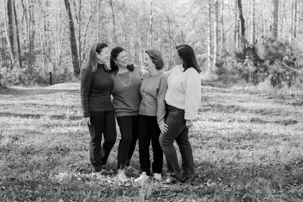 Black-and-white portrait of four women arm in arm, laughing together in a sunlit wooded clearing