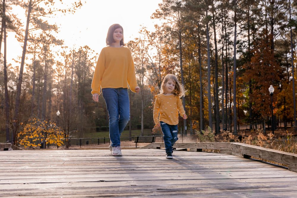 Two sisters in matching mustard sweaters and jeans walking across a wooden bridge in an autumn park