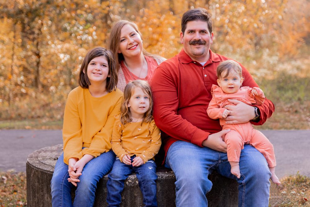 Family of five seated on a stone ledge in front of golden autumn foliage, father holding the baby