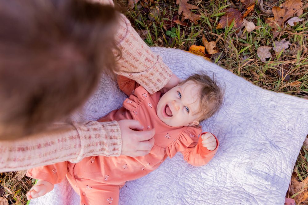 Overhead view of a mother tickling her laughing baby girl in coral lying on a quilt among fall leaves