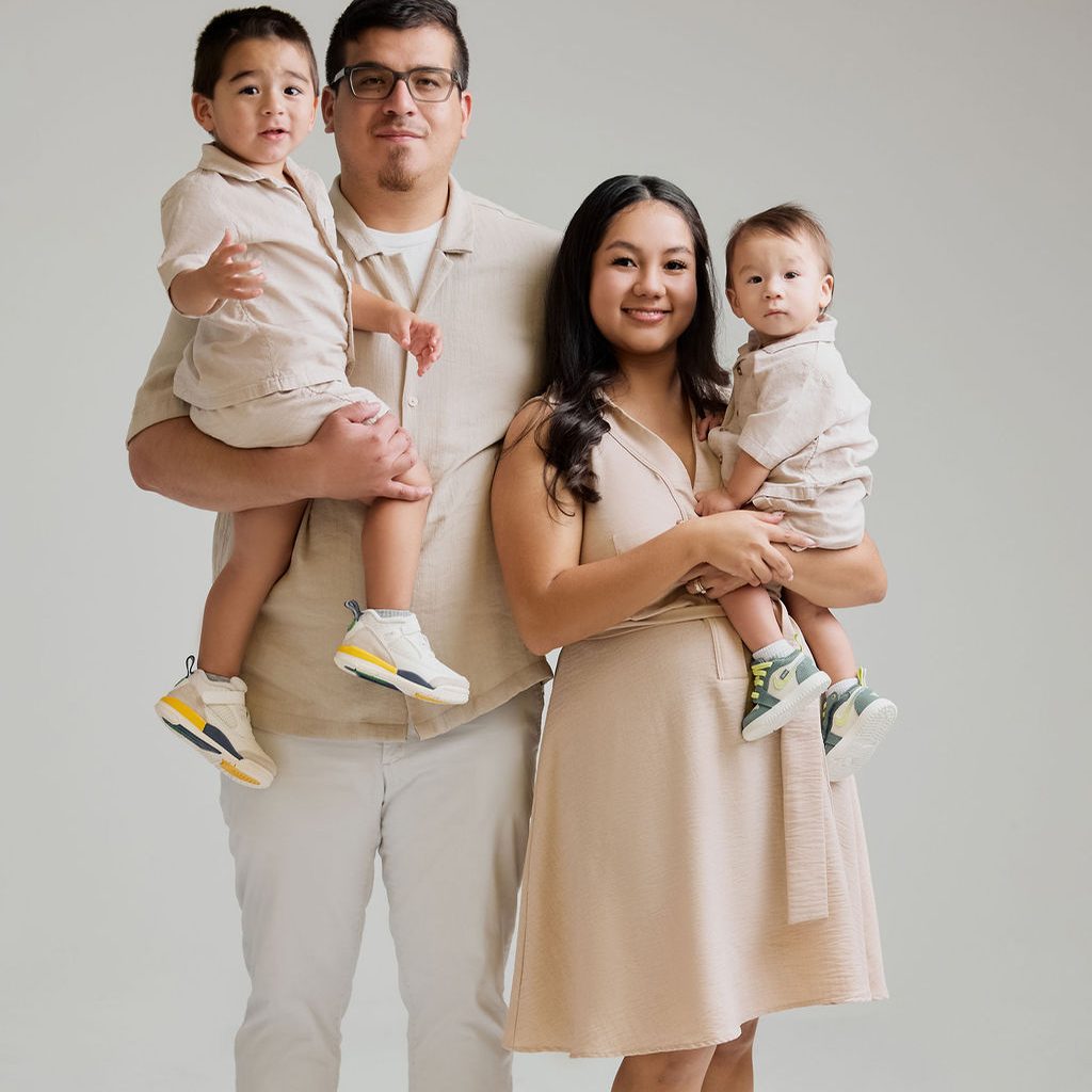 Family photography: parents in matching beige outfits each holding a toddler son against a light grey studio backdrop