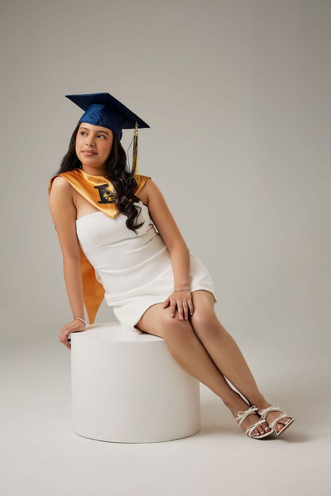 Graduate in a navy cap and gold stole sitting on a white cylinder pedestal, glancing over her shoulder