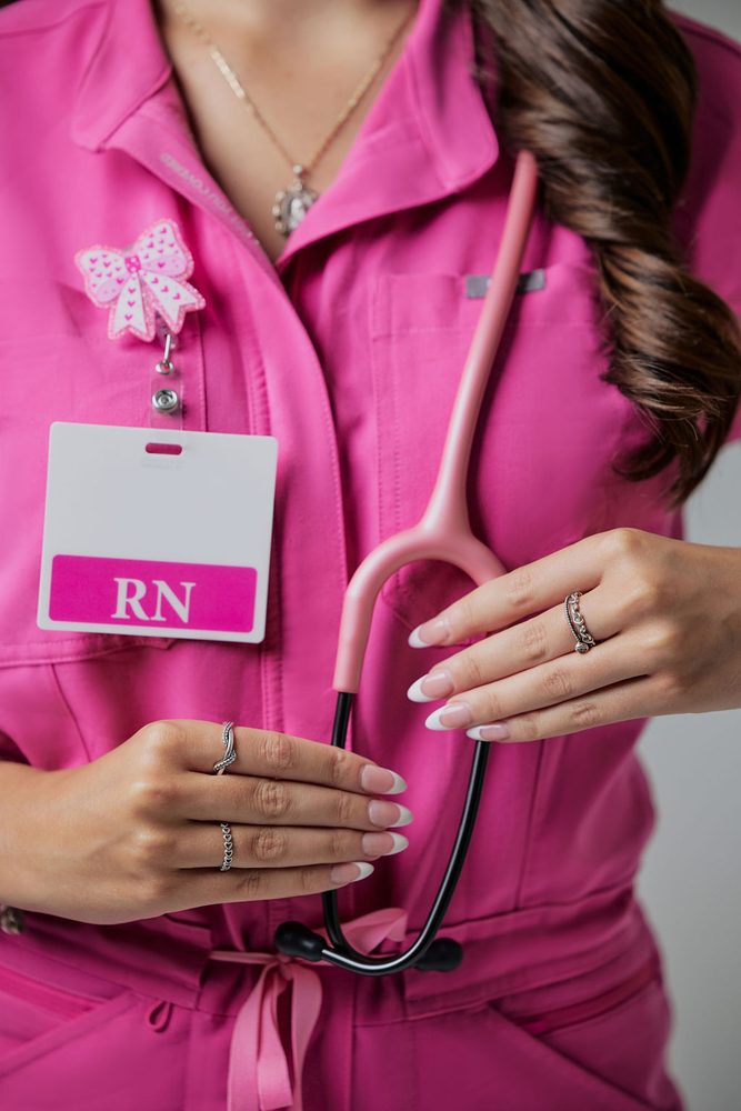 Close-up of a nursing graduate's hands holding a pink stethoscope beside an RN badge on pink scrubs