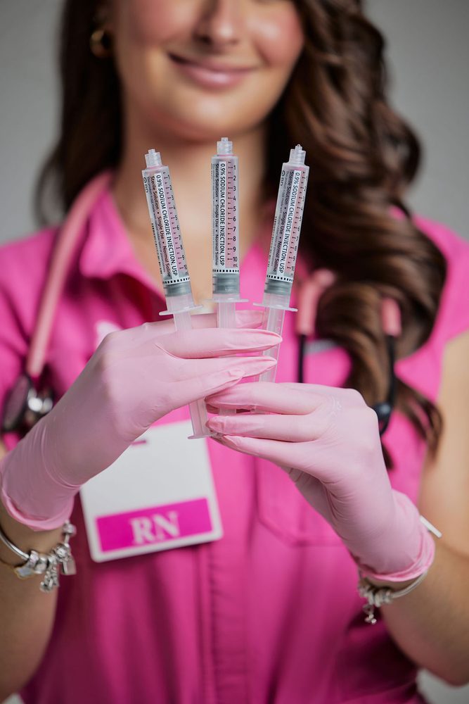Nursing graduate in pink gloves holding up three saline syringes, her RN badge softly blurred