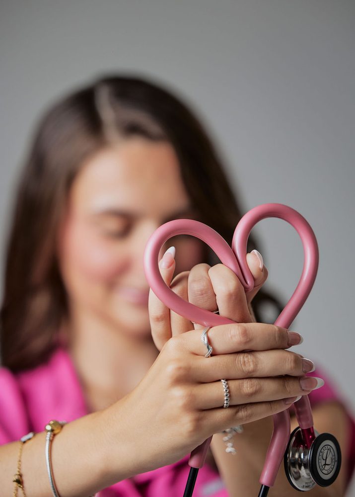 Nursing graduate shaping her pink stethoscope tubing into a heart, her face softly blurred behind