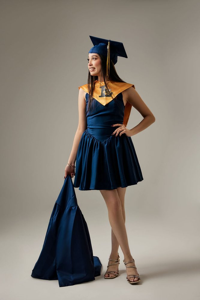 Graduate in a navy cap, gold stole and navy dress with a hand on her hip, holding her gown beside her