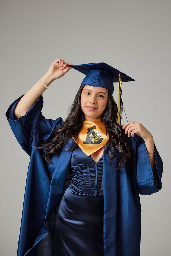Graduate in a navy cap and gown with a gold mascot stole, touching the brim of her cap and smiling softly