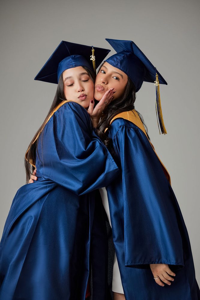 Two graduates in navy caps and gowns hugging cheek to cheek and making playful kissy faces