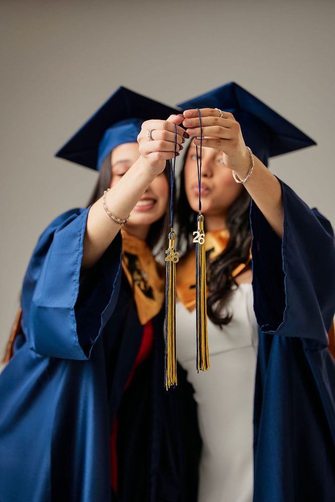 Two graduates in navy gowns holding up their gold-and-navy tassels with '26 charms toward the camera