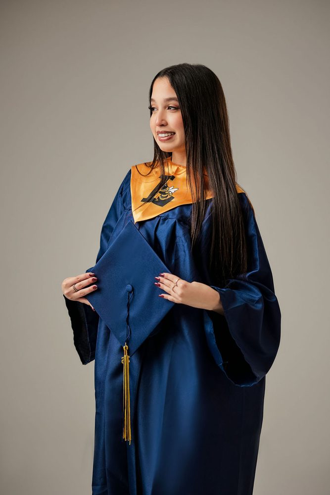 Smiling graduate in a navy gown and gold stole holding her mortarboard and tassel at her waist