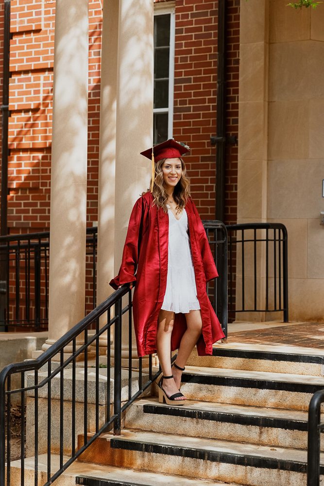 Graduate in a red cap and open gown over a white dress standing on stone steps by brick columns