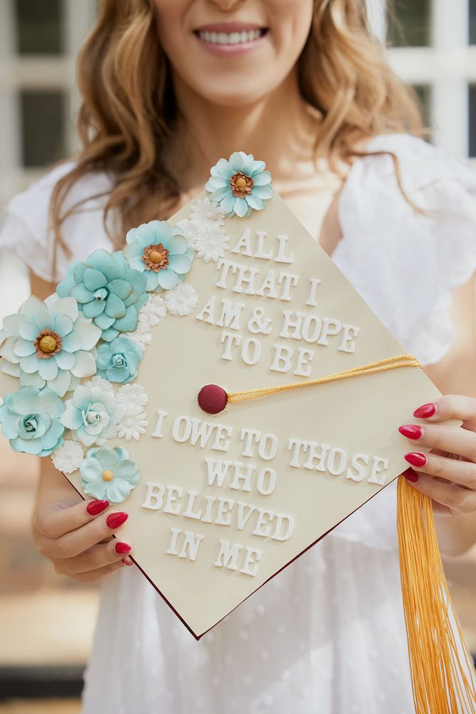 Graduate holding a cap decorated with teal paper flowers and a quote thanking those who believed in her