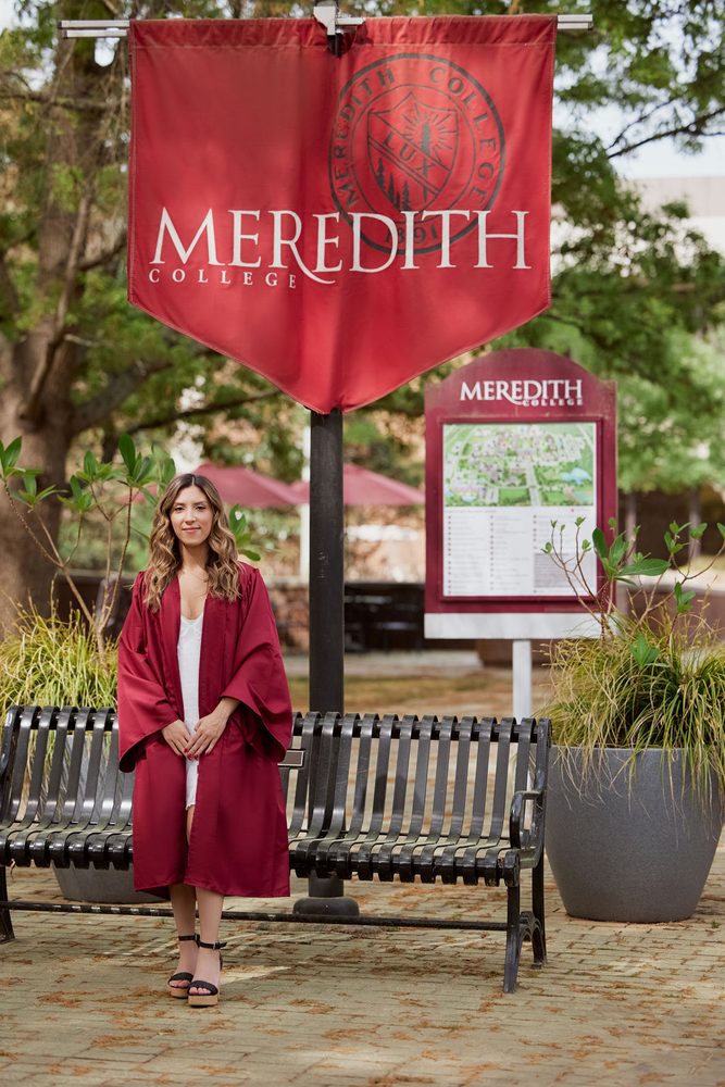 Graduate in a crimson gown standing by a bench beneath a Meredith College campus banner