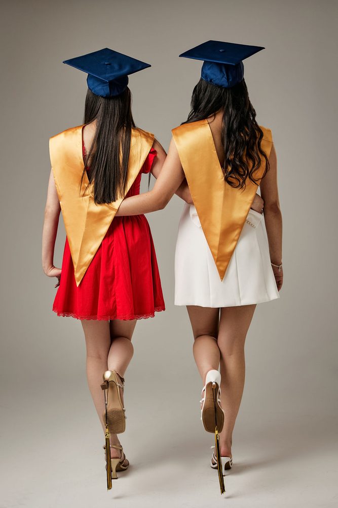 Back view of two graduates arm in arm in navy caps and gold stoles, one in a red dress and one in white