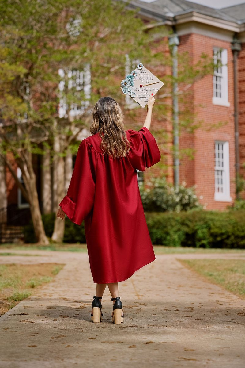Graduation photography: graduate in a red gown seen from behind, raising a flower-decorated cap on a campus walkway