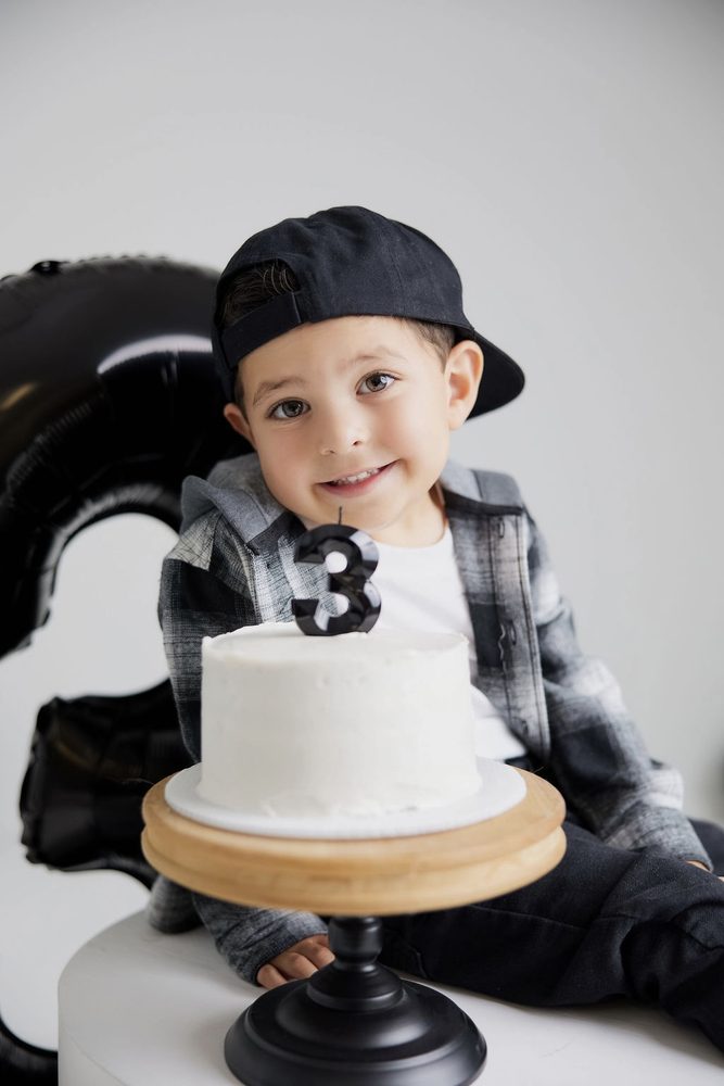 Little boy in a backwards cap smiling behind a white birthday cake with a black '3' candle on a wooden stand