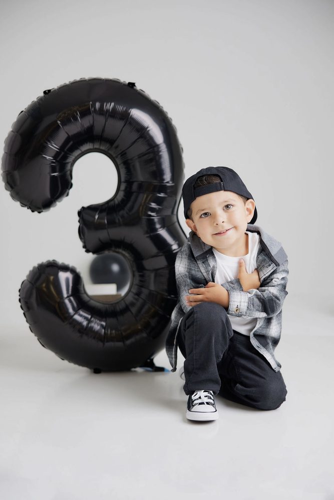 Boy in plaid flannel and backwards cap kneeling with a hand on his heart next to a giant black '3' balloon